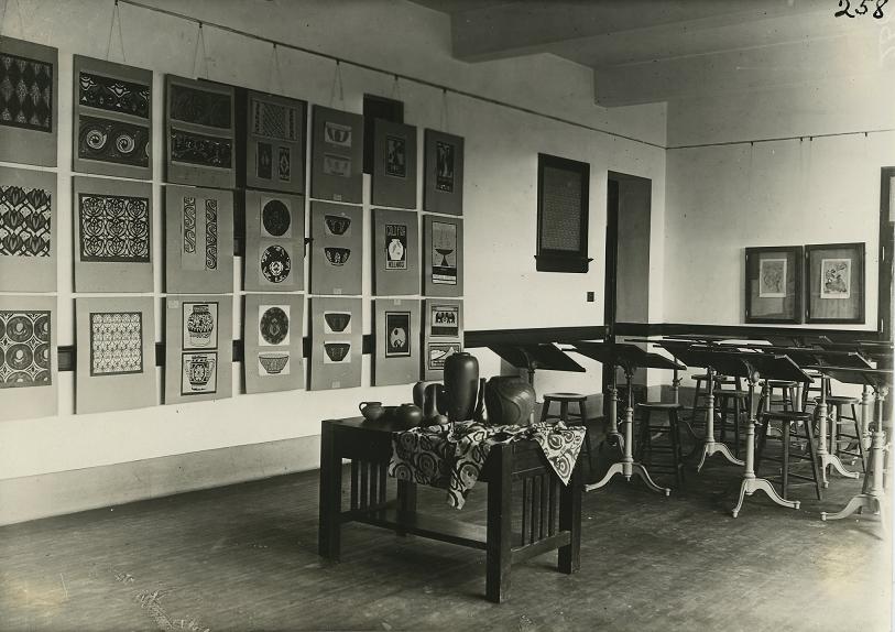 Art classroom located in Old Main. Walls adorned with various peices of art featuring pottery and pottery patterns. A table with a towel and pots in front of empty chairs and stands.